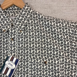May include: A cream-coloured button-down shirt featuring a black and grey paisley pattern. The shirt has a pocket and a tag that reads "No Wrinkle Prints Friday Shirt Bugle Boy Co."