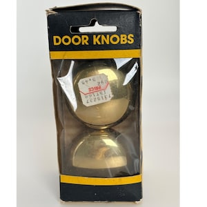 May include: A pair of round, polished brass door knobs packaged in a rectangular cardboard box with a clear plastic window. The black and yellow packaging features the text DOOR KNOBS at the top and a white price sticker on the top knob.