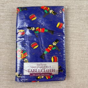 May include: A blue vinyl flannel-backed tablecloth featuring a repeating pattern of toy soldiers playing instruments and red drums. The packaging label reads HomeTrends Vinyl Flannel Back TABLECLOTH 60" x 84" OBLONG in black text.