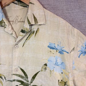 May include: A light tan button-down shirt with a floral pattern of blue flowers and green leaves. The shirt has the brand name "Tommy Bahama" on the collar. The shirt is hanging on a wooden hanger.