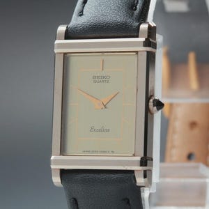 May include: A rectangular Seiko Quartz Exceline watch with a silver-tone case and a black leather band. The watch face is a light champagne color with gold-tone hands and markers. The watch has the text "SEIKO QUARTZ" and "Exceline" on the face.