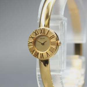 May include: A gold-tone Seiko wristwatch featuring a round dial with black Roman numeral hour markers on the bezel. The watch has a smooth, solid gold-tone bangle-style band and a small crown on the right side of the circular watch case.