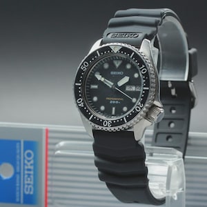 May include: A black Seiko dive watch with a black rubber strap. The watch has a black dial with white markers and hands. The bezel is black with white markings. The watch is displayed on a clear stand.