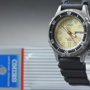 May include: A Seiko Professional 200m dive watch with a black bezel and black rubber strap. The watch has a cream-colored face with luminous hands and markers. The date display shows "FRI 14". The watch is displayed on a clear stand.