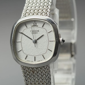 May include: A silver Credor Seiko wristwatch with a textured white face and silver mesh band. The watch has a rectangular face with black hands and hour markers. The watch is a classic design.