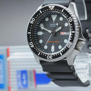 May include: A Seiko automatic diver's watch with a black dial, black rotating bezel, and black rubber strap. The watch face shows the day and date, with luminous markers. Water resistant to 200m. The watch has a stainless steel case.