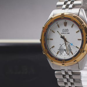 Puede incluir: Reloj Alba Quartz World Time plateado y dorado con esfera plateada y correa de acero inoxidable. El reloj tiene un bisel dorado y una esfera más pequeña. El reloj se exhibe sobre un soporte transparente.
