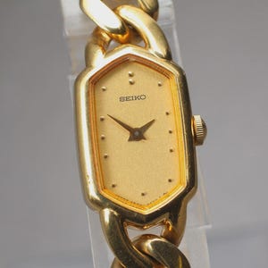 May include: A gold-toned Seiko wristwatch with a rectangular face and a chain-link band. The watch face is gold with black hands and the Seiko logo. The band is a thick, textured chain.