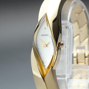 May include: A gold-tone Seiko wristwatch featuring a white, diamond-shaped dial with gold hands and a small gold dot at the twelve o'clock position. The watch has a polished, curved gold-tone metal band that integrates seamlessly with the case.
