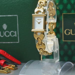 May include: A gold-toned Gucci watch with a white face and a gold chain-link bracelet. The watch is displayed on a clear stand, with a green Gucci box in the background. Additional watch straps are visible.