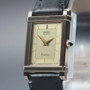 May include: A rectangular Seiko Quartz Exceline watch with a gold-tone face and hands, set in a gold-tone case. The watch features a black leather band. The watch face displays the brand name and model.