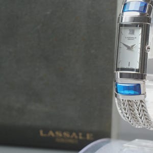 May include: A silver Lassale Seiko watch with a rectangular face and a woven metal band. The watch features blue accents on the top and bottom of the face. The watch is displayed on a clear stand.