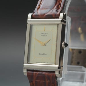 May include: A rectangular Seiko Quartz Exceline watch with a cream-colored face, gold hands, and a brown leather strap. The watch has a silver-colored case and a black crown. The watch face displays the Seiko Quartz logo and the word "Exceline".