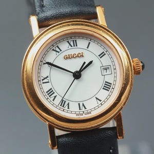 Puede incluir: Un reloj de pulsera dorado con caja redonda y esfera blanca con números romanos negros. La esfera muestra el logotipo de Gucci en letras doradas, manecillas negras y una ventana de fecha en la posición de las tres. Tiene una correa de cuero negro.