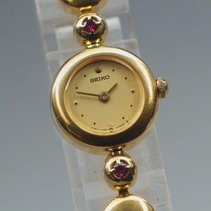 Puede incluir: Un reloj de pulsera Seiko dorado con esfera redonda y correa dorada. La esfera es de color crema con agujas e indicadores de hora dorados. La correa presenta detalles de color rubí. El reloj se exhibe sobre un soporte transparente.