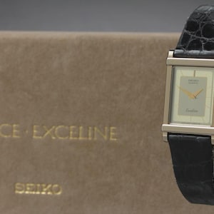May include: A rectangular Seiko Dolce Exceline watch with a silver-tone case and a black leather band. The watch face is light beige with gold hands and the word "Exceline" printed on the face. The watch is displayed on a clear stand.