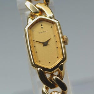 May include: A gold-toned Seiko watch with a rectangular face and a matching chain-link band. The watch face is a light gold color with black hands and hour markers. The watch has a classic, elegant design.