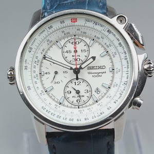 Op de afbeelding: Een Seiko chronograaf horloge met een witte wijzerplaat, zilverkleurige wijzers en uurmarkeringen. De wijzerplaat heeft drie sub-wijzerplaten, een rekenliniaalring met numerieke markeringen en een blauwe, gestructureerde lederen band. Tekst Seiko, Chronograph, 100M.