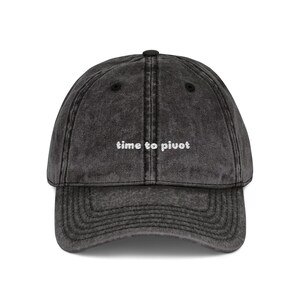 May include: A dark grey baseball cap with a curved peak. The cap has the embroidered text "time to pivot" in white, centred on the front. The hat has a vintage, worn appearance.