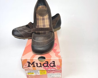 May include: A pair of dark brown Mary Jane shoes with a textured finish, a single strap across the instep, and a plaid interior lining. The shoes are displayed on top of a pink and orange Mudd brand shoebox with visible product labels.