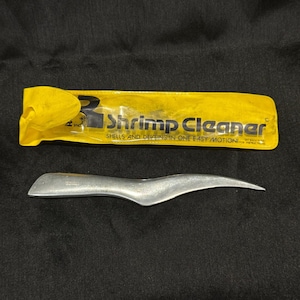 May include: A silver-colored metal shrimp cleaner tool with a curved, pointed blade, positioned below its original yellow plastic storage sleeve which features the black printed text Shrimp Cleaner Shells and Deveins in One Easy Motion.