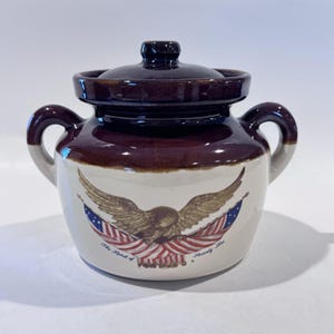 May include: A brown and cream-colored ceramic crock with a lid and two handles. The crock features a design of an eagle with outstretched wings, American flags, and the text "The Spirit of Seventy Six."