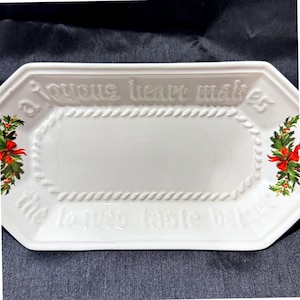 May include: A white rectangular ceramic serving tray with beveled corners. The rim features a raised rope texture. Embossed text reads: a joyous heart makes the loaves taste better. Holly and red ribbon motifs decorate the left and right sides.
