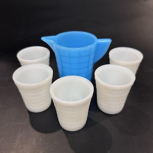May include: A blue pitcher with a handle and a spout, surrounded by five white, ribbed shot glasses. The pitcher and glasses are made of glass. The shot glasses are arranged around the pitcher.