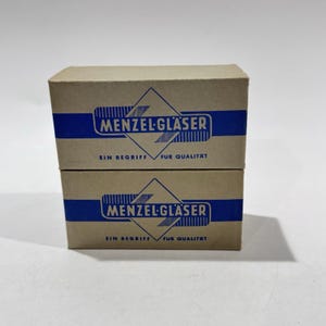 May include: Two stacked cardboard boxes with blue and white text. The boxes have a brown background with a blue stripe across the top. The text on the boxes reads "MENZEL-GLÄSER" and "EIN BEGRIFF FÜR QUALITÄT".