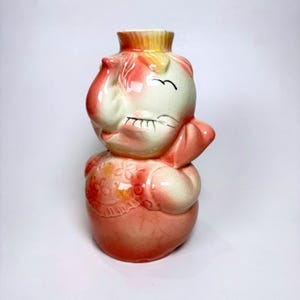 May include: A ceramic vase shaped like a stylized elephant with a cream and coral-pink gradient glaze. The elephant features a trunk, closed eyes with painted lashes, and a textured floral pattern on its chest, finished with a crackle glaze.