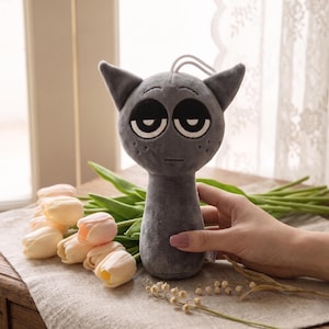 Puede incluir: Un juguete de peluche gris con forma de gato con ojos grandes y somnolientos y orejas puntiagudas. El juguete tiene un lazo de cuerda en la parte superior. El fondo incluye flores y una ventana.
