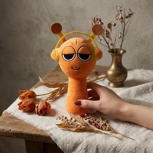 Könnte beinhalten: Ein orangefarbenes Plüschtier in Form einer Zeichentrickfigur mit großen Augen und Kopfhörern. Das Spielzeug hat braune antennenartige Merkmale und ein lächelndes Gesicht. Das Spielzeug wird von einer Person gehalten. Der Hintergrund umfasst getrocknete Blumen, eine Messingvase und einen Holztisch.