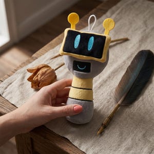 May include: A plush robot toy with a black screen face, blue eyes, and yellow antenna. The robot has a gray body with a yellow and gray striped neck. A dried flower and a feather quill are also in the image.