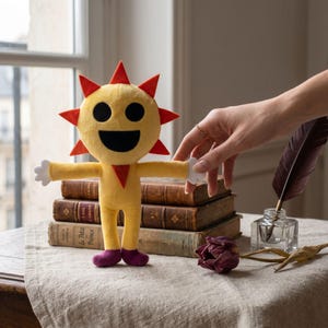 Puede incluir: Un peluche amarillo con forma de sol sonriente, con púas rojas, ojos negros y una boca ancha. Tiene los brazos extendidos con manos blancas, cuerpo amarillo y zapatos morados. El juguete está sobre una mesa con libros.