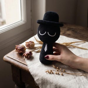 Könnte beinhalten: Schwarze Plüschpuppe mit Zylinder, weißer Krawatte und geschwungenen weißen Augenbrauen. Die Puppe wird von einer Person mit rotem Nagellack gehalten. Trockenblumen und ein Fenster im Hintergrund.
