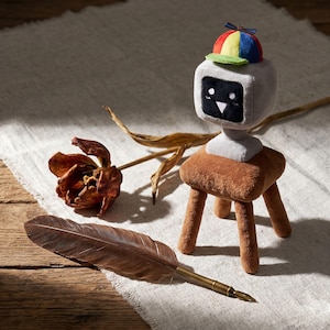 May include: A soft toy robot wearing a colourful cap sits on a small brown stool. A dried tulip and a quill pen with a bronze nib are also in the image. The background is a light-coloured fabric on a wooden surface.