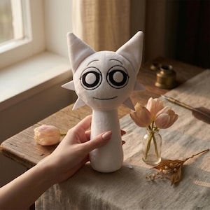 Può includere: Un peluche bianco con un volto simile a un gatto, con grandi occhi neri e una bocca sorridente. Il giocattolo è tenuto in mano, con un tavolo di legno e fiori sullo sfondo. Il giocattolo ha orecchie a punta e un design a forma di stella.