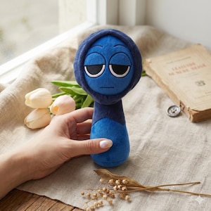 Puede incluir: Una muñeca de peluche azul con una cabeza con capucha y una expresión triste. La muñeca tiene ojos grandes y ovalados y una boca pequeña. La muñeca está sujeta por una mano, con flores y un libro en el fondo.