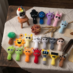 May include: A selection of colourful plush toys with diverse character designs. The toys feature various shapes, including suns, mushrooms, and abstract figures. The colour palette includes shades of green, yellow, orange, red, blue, and grey.