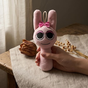 May include: A pink plush bunny toy with large black eyes, a pink bow, and a smiling face. The bunny has long ears and a loop for hanging. The toy is held in a hand, with a dried flower and a wooden table in the background.