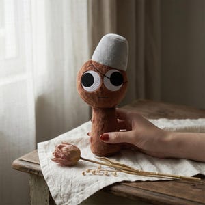 May include: A brown plush doll with large, round eyes and a gray hat. The doll is held by a hand with red nail polish. A dried tulip and sprigs of dried flowers rest on a white cloth.