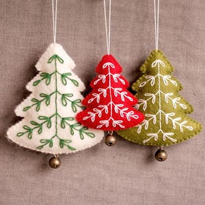 Puede incluir: Tres adornos de fieltro para árbol de Navidad en blanco, rojo y verde, cada uno con detalles bordados en blanco y una pequeña campana dorada. Los adornos están suspendidos por cuerdas blancas sobre un fondo neutro.