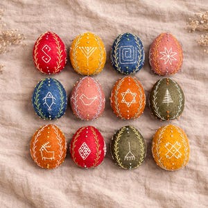 Könnte beinhalten: Eine Sammlung von zwölf bunten, bestickten Ostereiern. Jedes Ei hat eine andere Farbe, darunter Rot, Gelb, Blau und Orange, und weist ein einzigartiges weißes Stickmuster auf. Die Eier sind auf einem neutralfarbenen Stoff angeordnet.