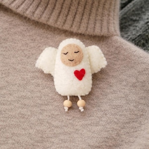 May include: A white felt angel pin with a red heart, beige face, and small wings. The angel has wooden bead legs and is pinned to a gray sweater. A charming, handmade accessory.