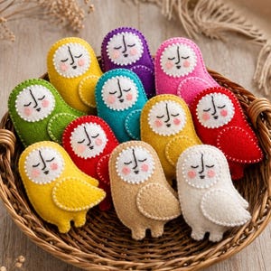 May include: A collection of eleven small, felt owl-shaped ornaments with embroidered faces, closed eyes, and pink cheeks. Each owl features a white facial disc and a solid-colored body in yellow, purple, pink, green, teal, red, tan, or white.