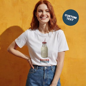 May include: White t-shirt with a graphic of a milk bottle with a red and white straw. The text "RAISED IN SHEFFIELD" is printed below the bottle in a red and blue rectangle. The background is a solid yellow-orange color.