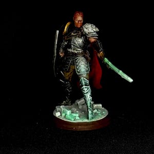 Puede incluir: Figura en miniatura de un guerrero con armadura plateada, empuñando una espada verde brillante y un escudo. El guerrero tiene barba roja y está sobre una base marrón. El fondo es negro.