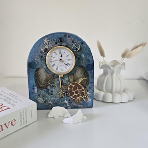 May include: A decorative clock with a blue resin arch, featuring a gold-rimmed clock face, seashells, and a turtle design. The clock is adorned with small anchors and starfish. A white vase with dried flowers and small white sculptures are also present.