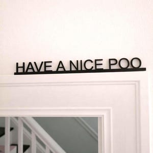 Op de afbeelding: Zwart houten bord met de tekst "HAVE A NICE POO" boven een witte deur. De tekst is in een eenvoudige, schreefloze lettertype en de letters zijn verbonden door een dunne balk.
