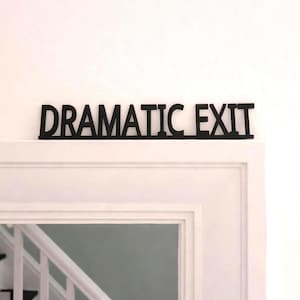 May include: Black wooden sign with the words "DRAMATIC EXIT" in bold, uppercase letters. The sign is mounted above a white door frame, creating a contrast between the dark text and the light background. The sign is a decorative element.